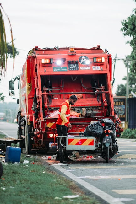 A middle-aged man with dark hair, wearing a black T-shirt with white text and dark trousers, is seen leaning forward and inserting a piece of rubbish into a silver metallic recycling bin on a paved si