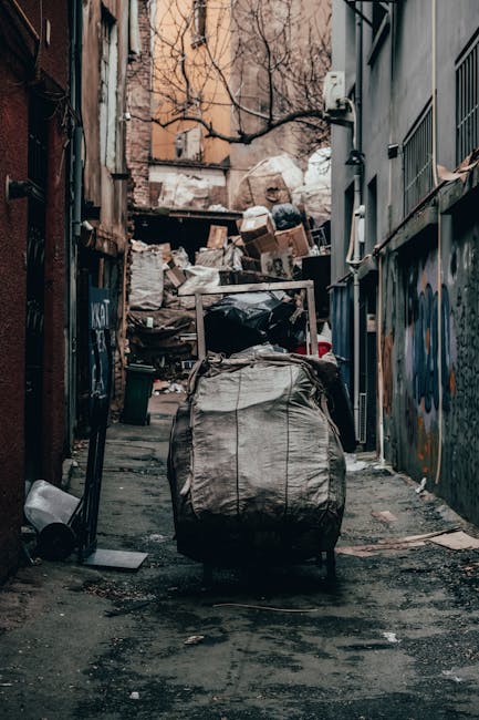 A narrow urban alleyway cluttered with a large, grey, wheeled rubbish bag in the foreground, positioned centrally on the damp, uneven pavement. The alley is enclosed by tall, worn brick and concrete b