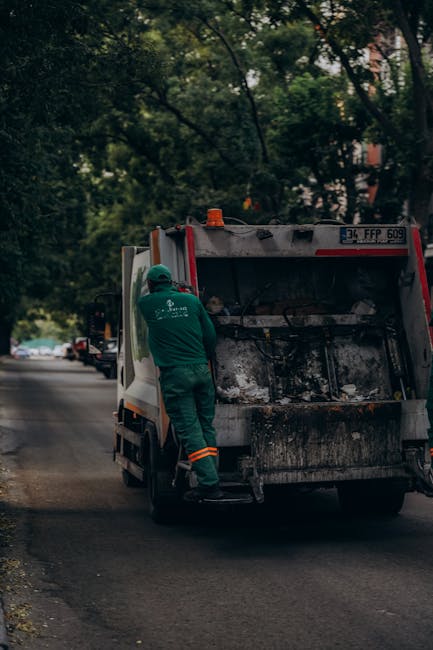 A waste collection vehicle parked on a narrow cobbled street in an urban area, with its hydraulic hopper open at the rear exposing rusty metal and mechanical components. A man wearing a blue uniform a
