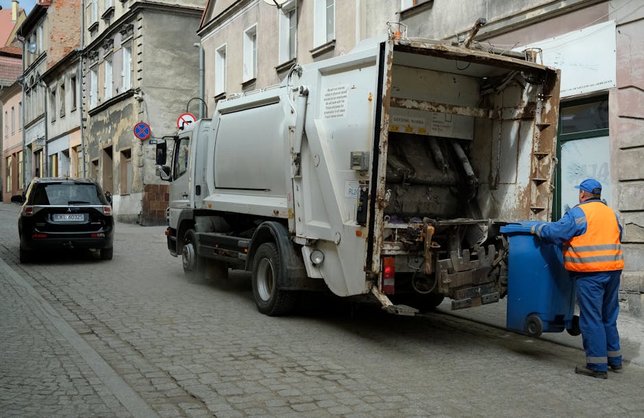 A small, dark red flatbed truck parked on a city street in front of a modern office building with large glass windows and concrete panels. The truck's cargo bed is filled with various types of rubbish